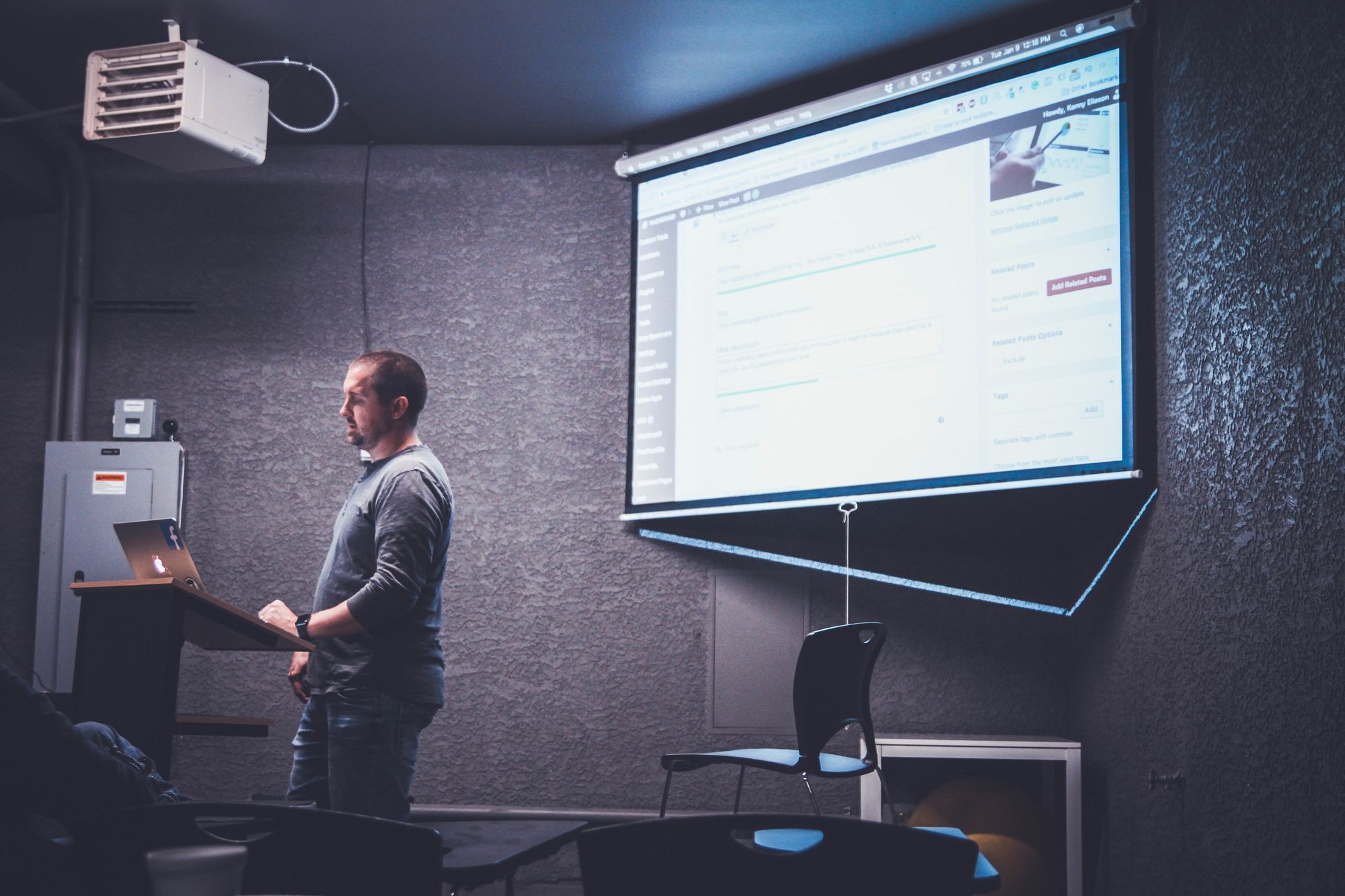 Stock image of a male-presenting person reading from a laptop, the screen of which is being projected on a white backdrop to present to a class of people. 