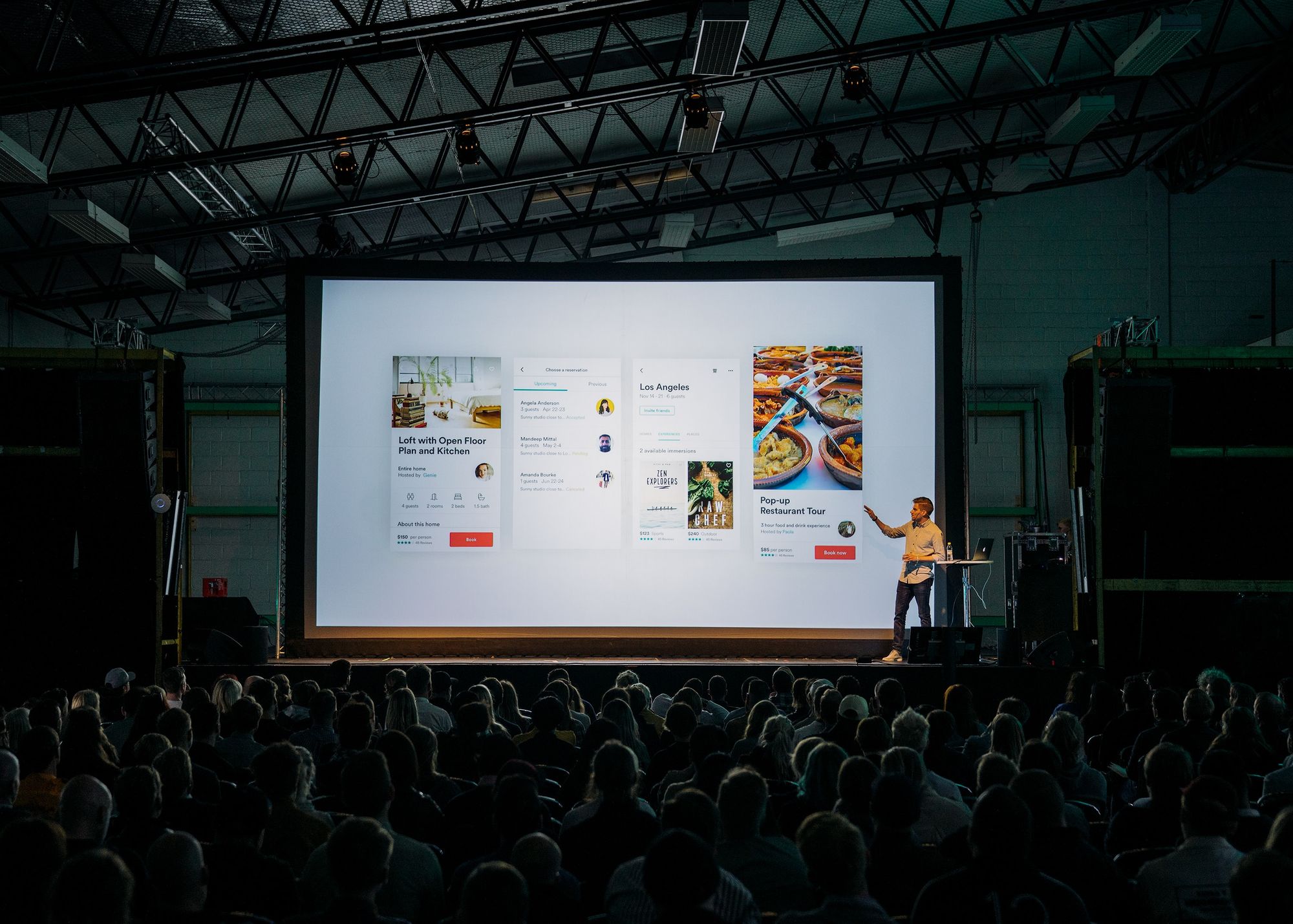 A male presenting person presenting to a large group of people - everyone is sat with their backs to the camera, staring at a huge white screen which shows reviews of a pop-up restaurant.